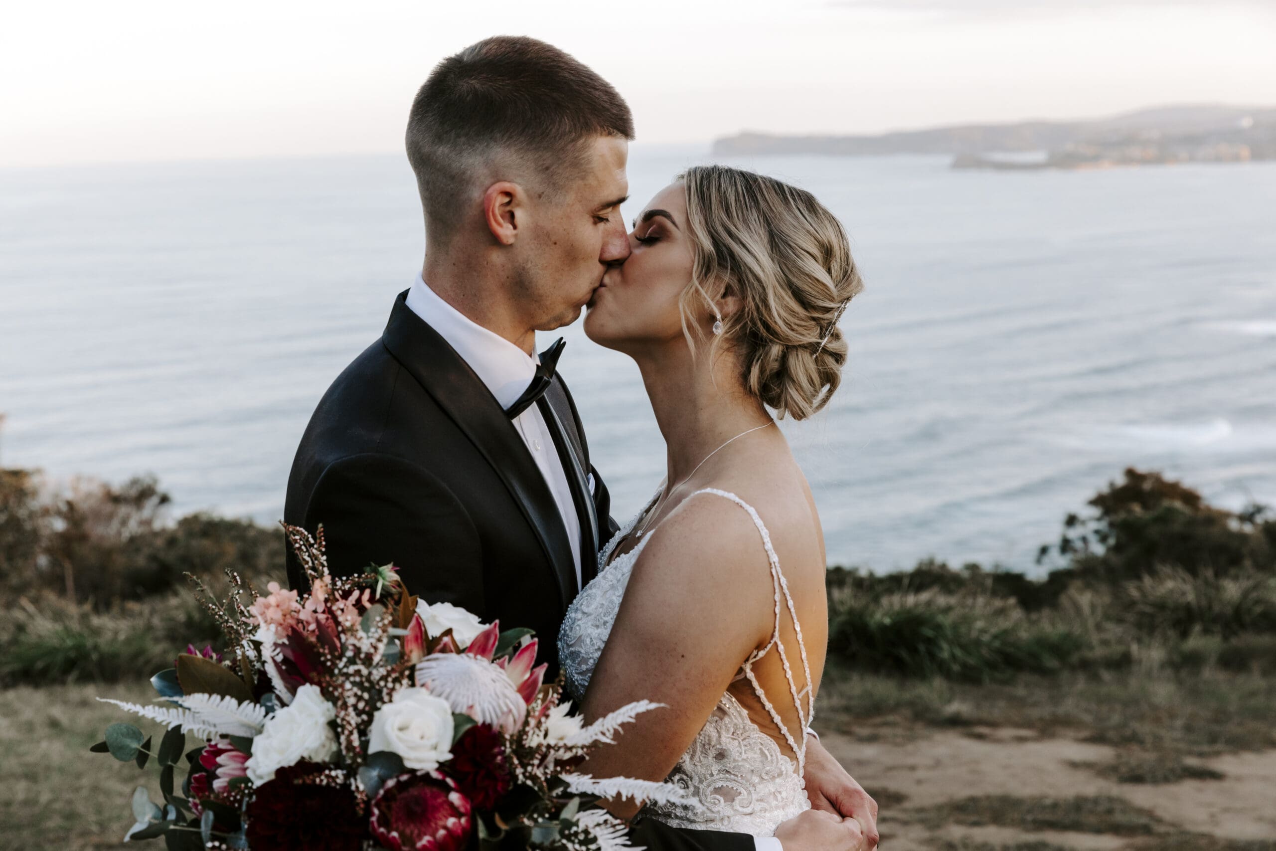 Bride and groom kissing at sunset while holding a bouquet with coastal views behind them.
