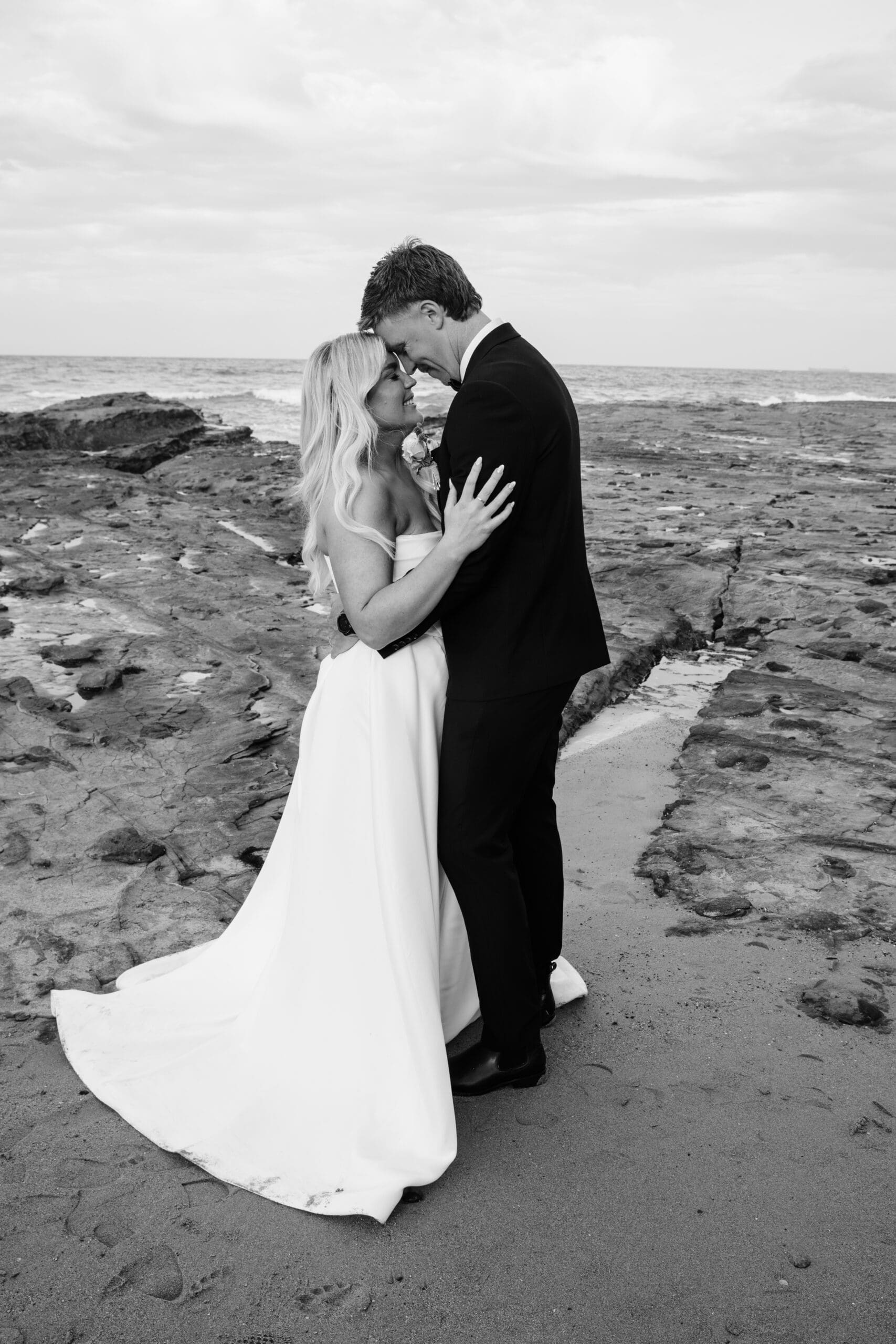 Bride and groom hugging on a rocky coastline during their wedding portraits.