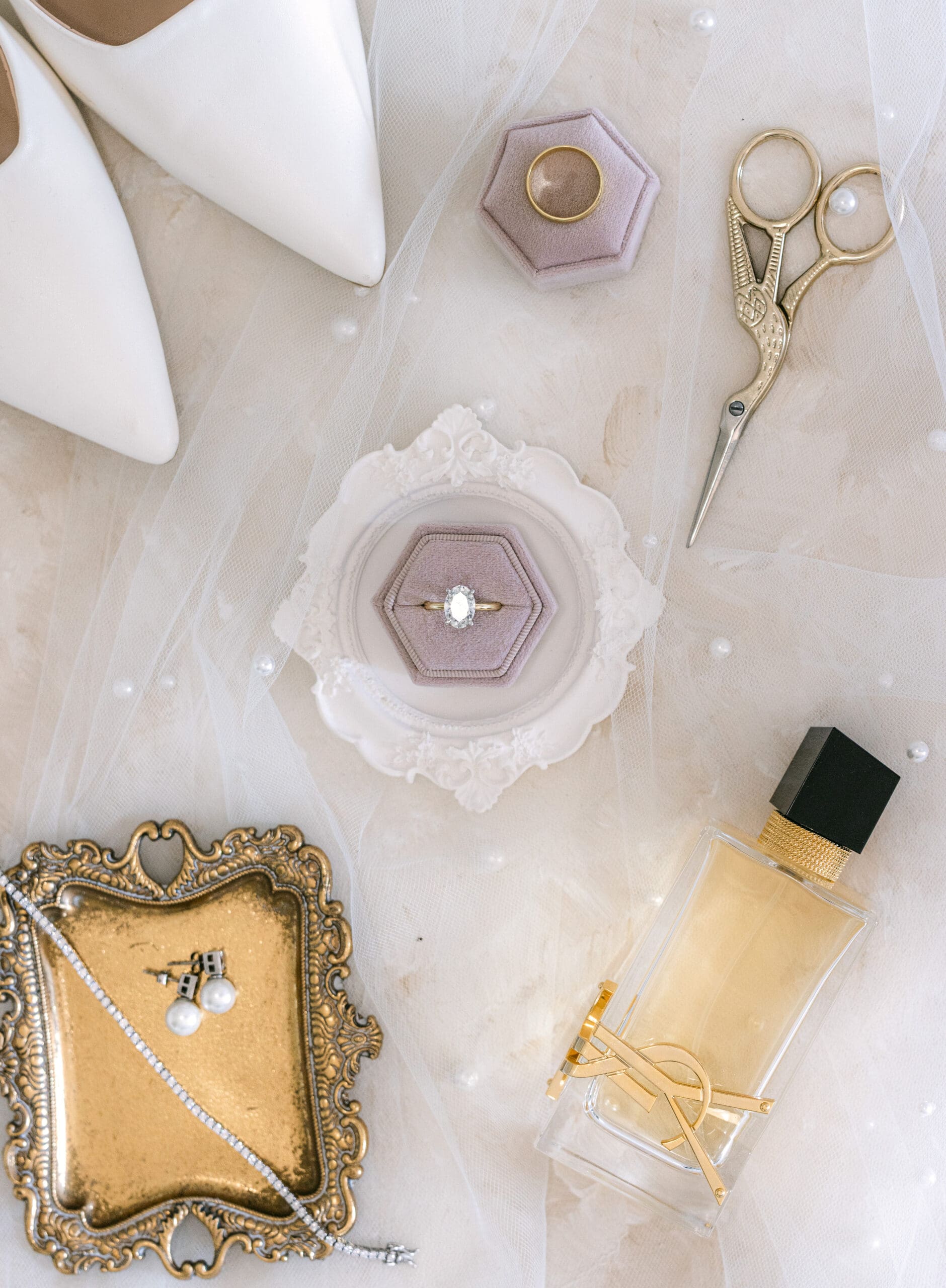 Flatlay of ring boxes, perfume, and gold tray on a soft white fabric background.
